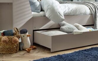 Closeup detail of the Cyclone Day Bed in Taupe from Pay Weekly Carpets, showing the pull-out drawer, which is supported beneath by casters to make it easy to move, and contains a mixture of cushions, books and soft furnishings as an indication of how much could be stored within. Also shown is a plush toy seagull and a basket containing books.