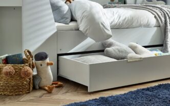 Close-up detail of the Cyclone Day Bed in White from Pay Weekly Carpets, showing a pull-out drawer supported by casters that contains some cushions and books to give an idea of the drawer's capacity.